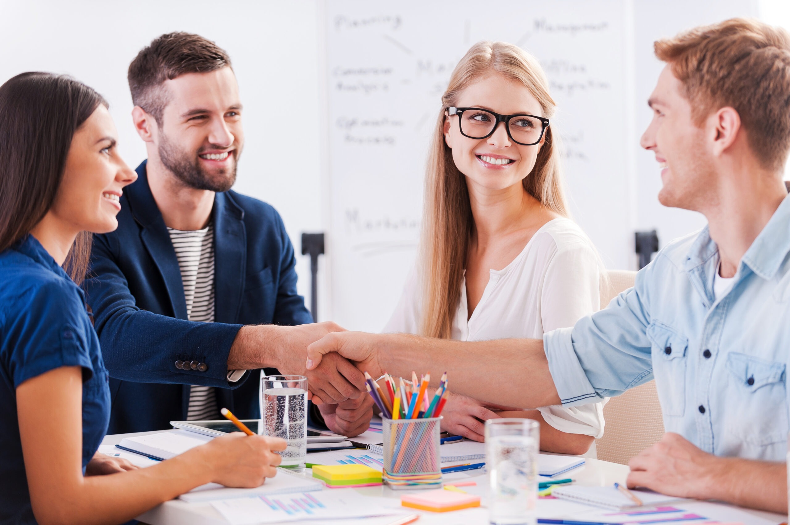 Group of cheerful business people sitting at the table together while two men shaking hands and smiling